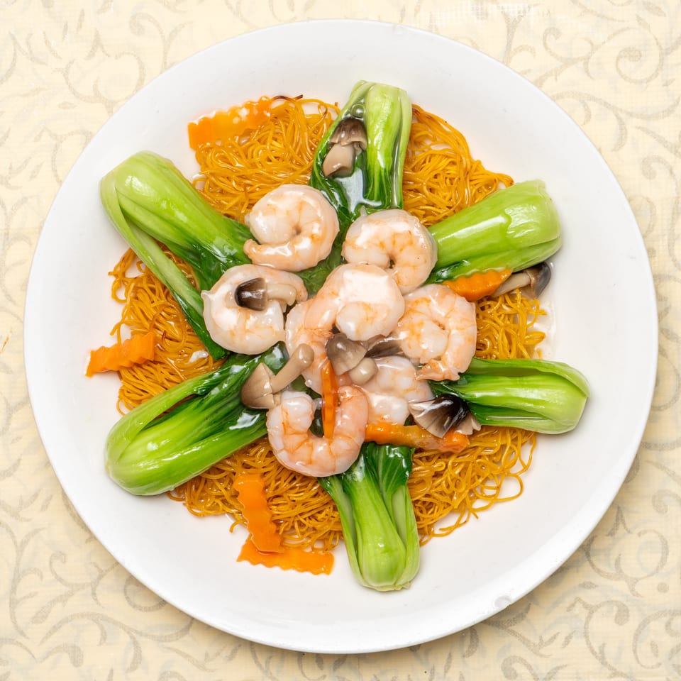 Crispy egg noodles topped with shrimp, bok choy, mushrooms, and carrots on a white plate.